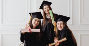 Three young graduates in gowns celebrate with diplomas indoors, exuding happiness.